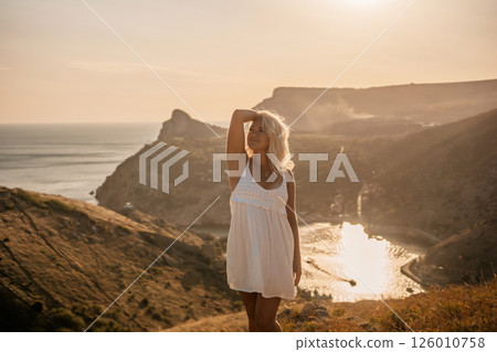 A blonde woman stands on a hill overlooking the ocean. She is wearing a white dress and she is enjoying the view. A blonde woman stands on a hill overlooking the ocean. She is wearing a white dress and she is enjoying the view. 126010758