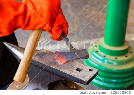 Skilled artisan shapes glowing axe making on an anvil while wearing protective gloves. 126010868