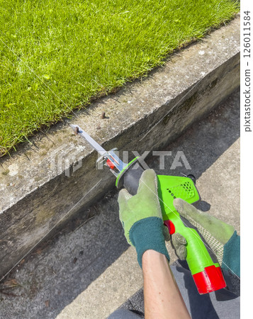 Male hands in green gloves cut a metal bolt protruding from concrete beam with electric reciprocating saw. Male hands in green gloves cut a metal bolt protruding from concrete beam with electric reciprocating saw. 126011584