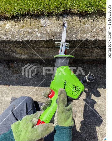 Male hands in gloves cutting a metal bolt protruding from a concrete beam with reciprocating electric saw. Close-up. 126011635