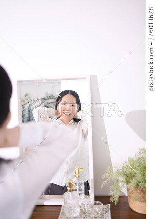 A smiling middle-aged woman putting on a necklace while looking in the mirror 126011678