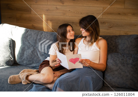 Mom and her preteen daughter reading together greeting card with drawn heart sitting on sofa at home. Spending quality time with children. 126011804