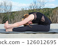 Woman practicing yoga outdoors on sandy surface, performing seated forward bend pose on mat, embracing relaxation and flexibility in natural surroundings under a clear sky. 126012695