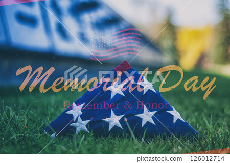A folded American flag rests on the grass near a military memorial wall, accompanied by the words Memorial Day Remember and Honor. A symbol of remembrance and national pride. 126012714