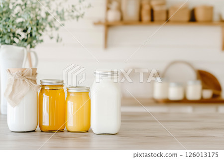 Three jars filled with a golden hue of honey and creamy white milk are artistically displayed on a light wooden kitchen surface. A subtle touch of greenery complements the cozy atmosphere. 126013175