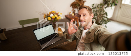 Portrait of young handsome man, cafe visitor taking selfie on smartphone app, showing okay, good recommendation sign, blogger rates coffee shop, nods in approval Portrait of young handsome man, cafe visitor taking selfie on smartphone app, showing okay, good recommendation sign, blogger rates coffee shop, nods in approval 126013755