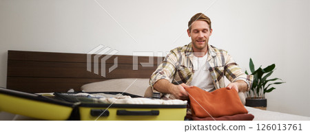 Portrait of handsome young man, tourist unpacking clothes in hotel room, staying in hostel on vacation, travelling, sitting with luggage on bed 126013761