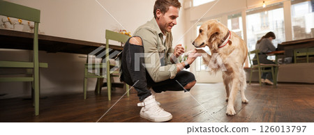 Close up portrait of smiling happy dog owner in pet-friendly cafe, teaching to give paw, giving a treat to his golden retriever, spending time in coffee shop 126013797