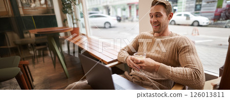 Portrait of handsome modern man, sitting in coffee shop with laptop, looking at screen with happy face, drinking cappuccino, resting in cafe or working 126013811