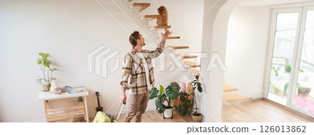 Vertical shot of smiling man with suitcase, playing with a cat, standing and carrying luggage Vertical shot of smiling man with suitcase, playing with a cat, standing and carrying luggage 126013862