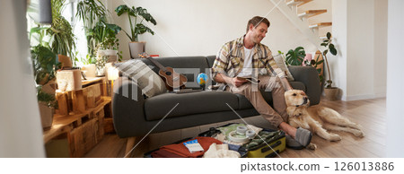 Man with a dog sitting near suitcase, holding notebook, checking list of items to take on vacation, planning a holiday trip Man with a dog sitting near suitcase, holding notebook, checking list of items to take on vacation, planning a holiday trip 126013886