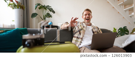 Happy man booking holiday online on laptop, paying for vacation on travel agency website, buying flight tickets in internet, sitting in living room with suitcase 126013903
