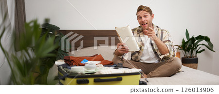 Portrait of happy, charismatic young man, excited about holiday, holding notebook, packing suitcase with clothes for vacation, pointing finger at camera and smiling Portrait of happy, charismatic young man, excited about holiday, holding notebook, packing suitcase with clothes for vacation, pointing finger at camera and smiling 126013906