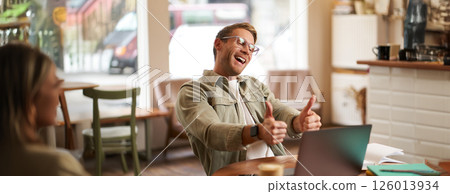 Portrait of satisfied, happy young man in glasses, working in cafe, sitting in co-working space with laptop, showing thumbs up, like and approve smth good, chatting, giving online lessons 126013934