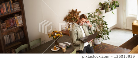 Image of young man, successful businessman sitting on table, looking at laptop, working in an office, looking concentrated on the project, doing job task, drinking coffee Image of young man, successful businessman sitting on table, looking at laptop, working in an office, looking concentrated on the project, doing job task, drinking coffee 126013939
