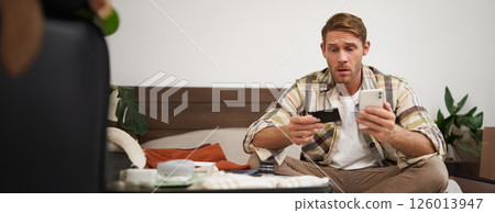 Portrait of young man, tourist paying online, using credit card and mobile phone app, sitting on bed with suitcase, ordering in internet, buying holiday tickets Portrait of young man, tourist paying online, using credit card and mobile phone app, sitting on bed with suitcase, ordering in internet, buying holiday tickets 126013947