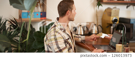 Portrait of young man, entrepreneur working from cafe, sitting with laptop, pressing his debit card to pay for order, prefer contactless payment, completing purchase Portrait of young man, entrepreneur working from cafe, sitting with laptop, pressing his debit card to pay for order, prefer contactless payment, completing purchase 126013973