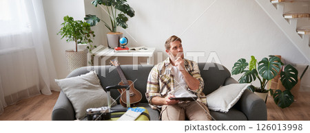 Image of young man with tablet, sitting in living room with suitcase, reading, studying the route, preparing for a vacation 126013998