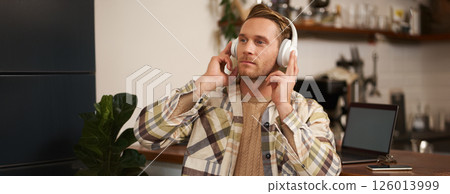 Portrait of handsome happy guy, freelancer working and listening to music in wireless headphones, dancing on his chair in cafe, enjoying favourite song in earphones 126013999