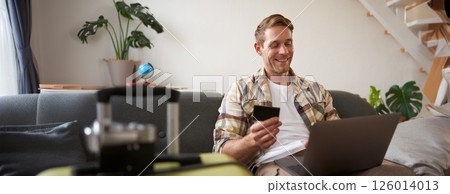 Happy man booking holiday online on laptop, paying for vacation on travel agency website, buying flight tickets in internet, sitting in living room with suitcase 126014013