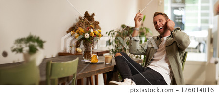 Happy handsome man in coffee shop, listening to music in wireless earphones, connects to cafe wifi to enjoy favourite song on his smartphone playlist, sitting and drinking cappuccino Happy handsome man in coffee shop, listening to music in wireless earphones, connects to cafe wifi to enjoy favourite song on his smartphone playlist, sitting and drinking cappuccino 126014016