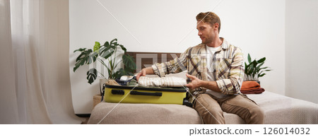 Portrait of young man packing his luggage for a trip, going on holiday, sitting on bed and putting clothes in suitcase, preparing for his flight Portrait of young man packing his luggage for a trip, going on holiday, sitting on bed and putting clothes in suitcase, preparing for his flight 126014032
