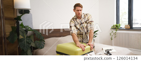Portrait of man sitting on bed, struggling to close full suitcase, packing for holiday, going on vacation Portrait of man sitting on bed, struggling to close full suitcase, packing for holiday, going on vacation 126014134