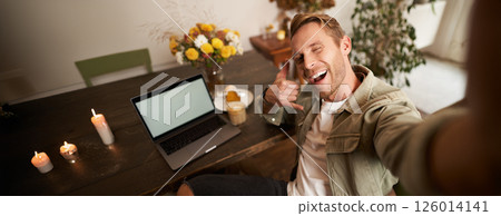 Handsome stylish young man takes selfie in cafe while working or studying remotely with laptop, shows call phone hand sign and smiling at mobile phone camera Handsome stylish young man takes selfie in cafe while working or studying remotely with laptop, shows call phone hand sign and smiling at mobile phone camera 126014141