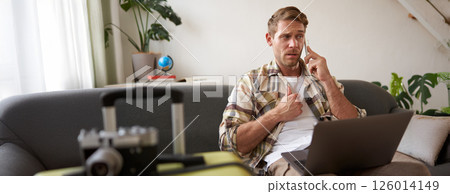 Man with confused face, talks on mobile phone, sits with laptop, has packed suitcase near him, enquires about holiday with travel agency 126014149