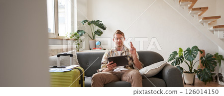 Man makes a wish, looks at digital tablet, has suitcase packed and ready for holiday, goes on vacation, picks tour on travel agency website 126014150