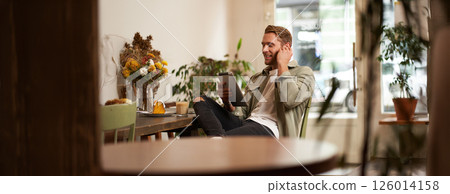 Portrait of handsome happy guy, young man sits in cafe, watching videos on digital tablet, wearing wireless headphones, laughing and smiling, spending time in coffee shop 126014158
