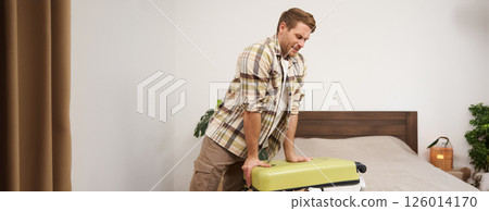 Portrait of young guy, traveller going on holiday, pressing his suitcase, trying to push clothes inside bag, closing his luggage, struggling to pack lots of things for vacation 126014170