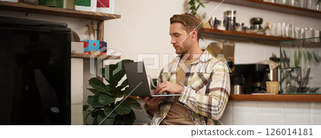 Image of young stylish guy with laptop, sitting in cafe with his computer, typing something, drinking coffee and doing project Image of young stylish guy with laptop, sitting in cafe with his computer, typing something, drinking coffee and doing project 126014181