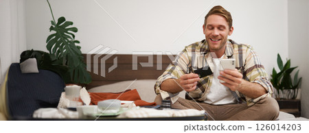Smiling tourist, man with suitcase, sits on bed, pays online, books a hotel, holds credit card and mobile phone, uses an app to purchase in internet Smiling tourist, man with suitcase, sits on bed, pays online, books a hotel, holds credit card and mobile phone, uses an app to purchase in internet 126014203