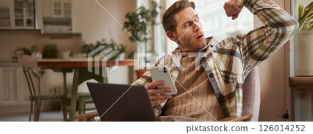 Portrait of young athletic guy sitting in cafe with laptop and smartphone, showing his biceps, flexing muscles and looking pleased at himself Portrait of young athletic guy sitting in cafe with laptop and smartphone, showing his biceps, flexing muscles and looking pleased at himself 126014252