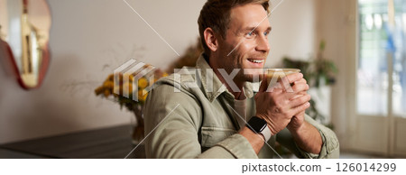 Leisure, people and lifestyle concept. Happy handsome young man in cafe, sitting on chair and smiling, holding glass of coffee, looking aside, relaxing with cup of cappuccino 126014299