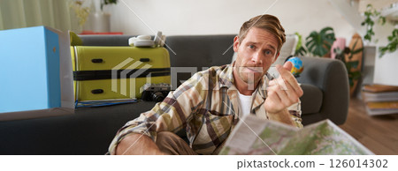Handsome smiling guy with road map, shows heart finger gesture, sits in living room with suitcase, plans holiday 126014302