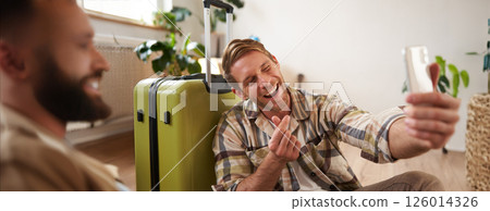 Smiling young man with suitcase, takes selfies on smartphone, blogger posts about his holiday tour on social media, using mobile phone Smiling young man with suitcase, takes selfies on smartphone, blogger posts about his holiday tour on social media, using mobile phone 126014326