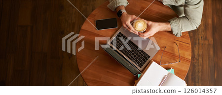 Top view. Male hands holding cup of coffee, man sitting at round table in a cafe, working on laptop, had his smartphone and notebook, drinking cappuccino 126014357