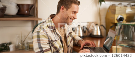 Portrait of handsome businessman, young digital nomad, sitting in cafe and looking at his laptop screen, working remotely, doing freelance project from coffee shop Portrait of handsome businessman, young digital nomad, sitting in cafe and looking at his laptop screen, working remotely, doing freelance project from coffee shop 126014423