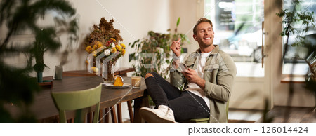 Portrait of young handsome man in casual clothes, sits in cafe and enjoys favourite song, listens to music in wireless headphones, vibing to the relaxing tunes in earphones, holding smartphone Portrait of young handsome man in casual clothes, sits in cafe and enjoys favourite song, listens to music in wireless headphones, vibing to the relaxing tunes in earphones, holding smartphone 126014424