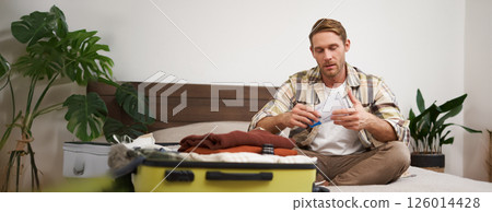 Portrait of handsome man, tourist, looking at his holiday tickets, booked vacation with travel agency, sitting on bed and packing clothes in suitcase 126014428