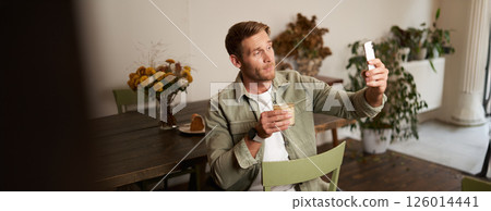 Portrait of young handsome man takes slefie on his mobile phone app, sits in cafe with glass of coffee, posting on social media application 126014441