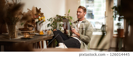 Portrait of young man sitting in cafe, spending time alone in coffee shop, reading on digital tablet, watching video, listening music in wireless headphones. Lifestyle and people concept 126014445