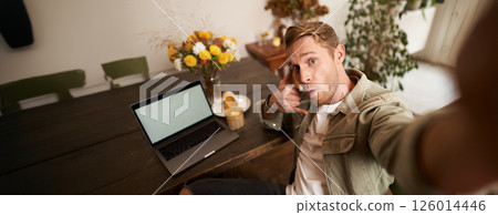 Handsome stylish young man takes selfie in cafe while working or studying remotely with laptop, shows call phone hand sign and smiling at mobile phone camera 126014446