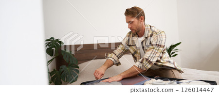 Portrait of young man, tourist going travelling abroad, putting clothes in suitcase, closing his luggage, sitting on bed, getting ready for holiday 126014447