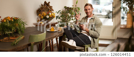 Portrait of happy handsome young man, sitting in cafe with mobile phone, wearing wireless headphones, listening to music or podcast while relaxing in coffee shop 126014451
