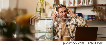 Lifestyle portrait of happy, excited young man, sitting in coffee shop with laptop and headphones, dancing on his chair and enjoying great quality sound, streaming favourite songs 126014505