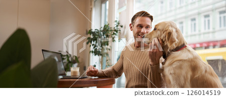 Lifestyle photo of young handsome man, entrepreneur working in a cafe on laptop while his dog sits next to him. A guy pets his golden retriever while using computer in a coffee shop Lifestyle photo of young handsome man, entrepreneur working in a cafe on laptop while his dog sits next to him. A guy pets his golden retriever while using computer in a coffee shop 126014519
