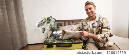 Portrait of young man sits on bed with suitcase, goes on holiday, packs clothes for vacation or business trip, checks his luggage 126014539